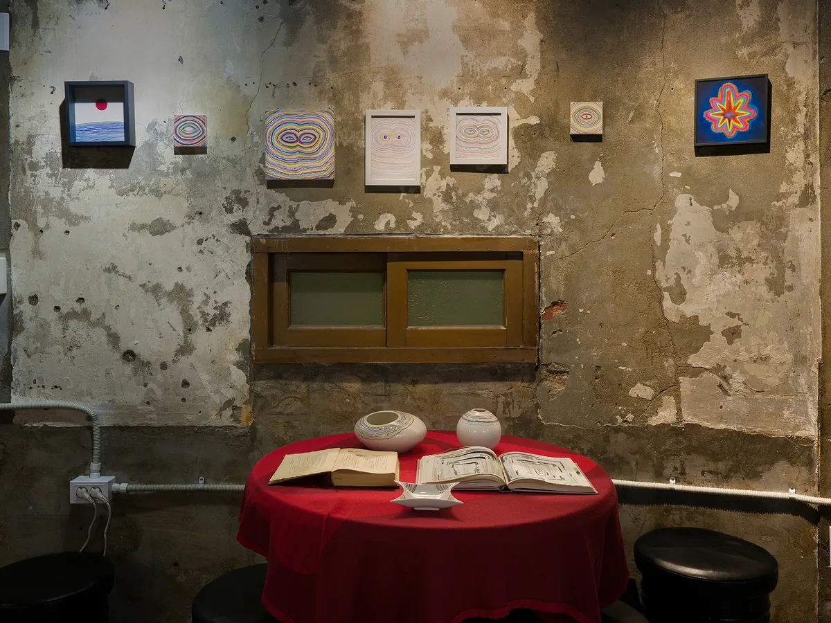Installation view — small works above a red table with ceramics and books